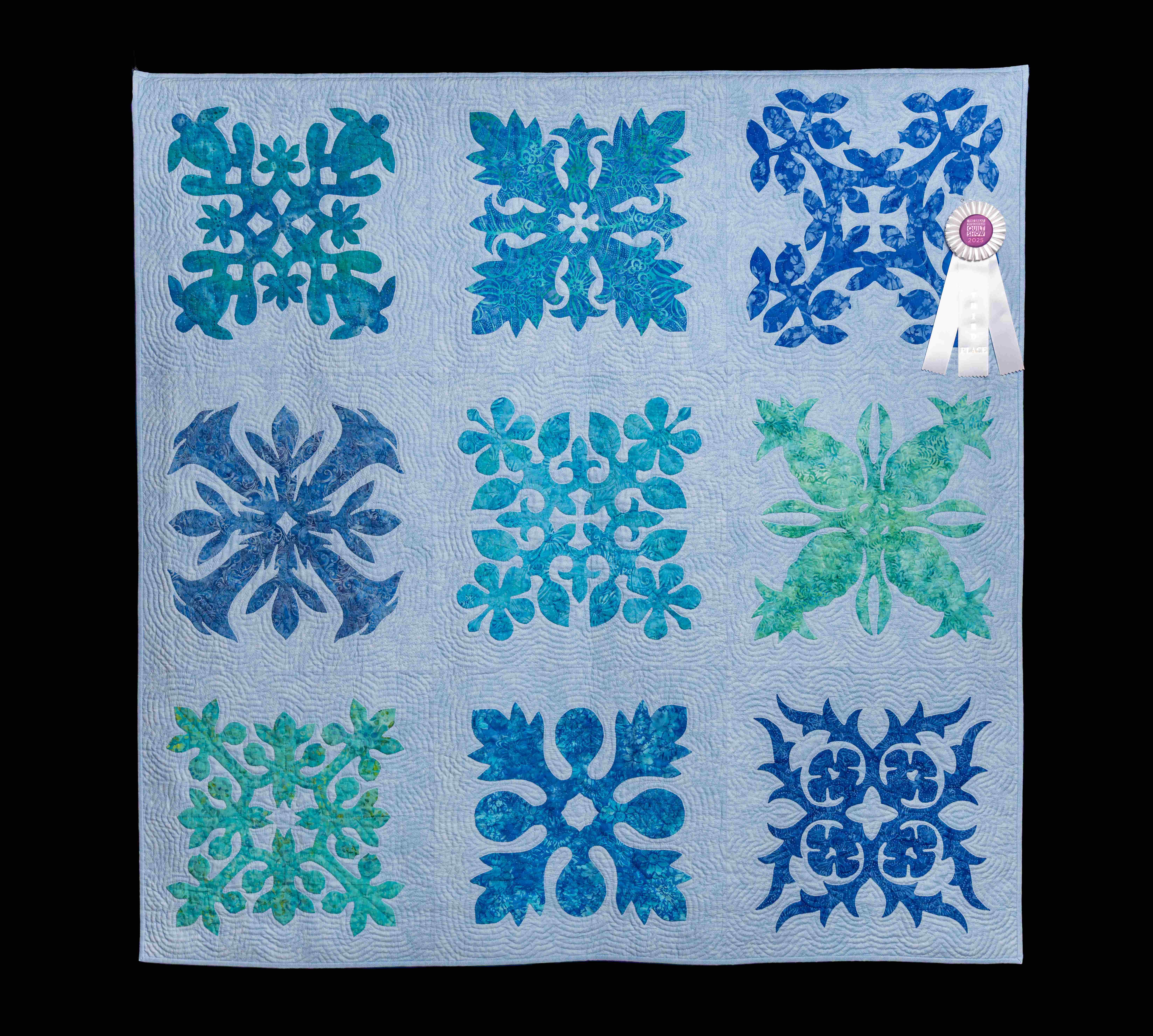 Award-winning quilt with nine symmetrical blue and green appliqué panels—botanical motifs on light blue, arranged in a 3×3 grid.