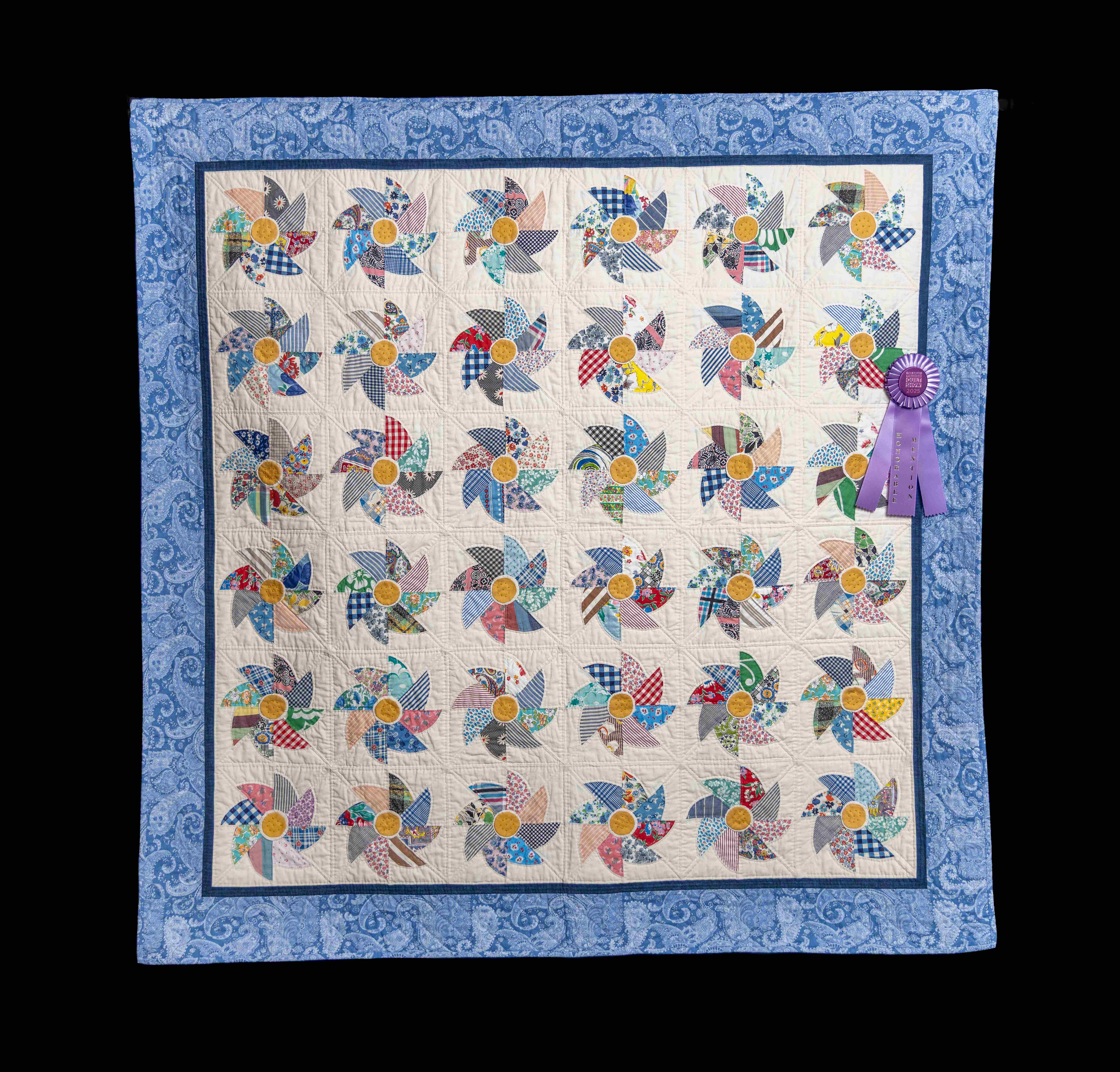 Award-winning quilt with colorful pinwheel blocks—yellow centers and patterned petals—set in a grid with a blue border and purple ribbon.